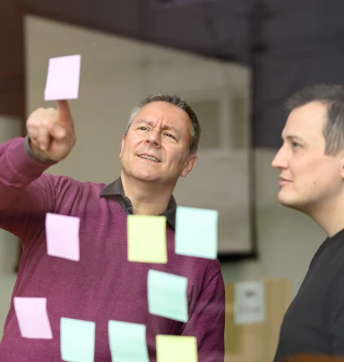 Jochen zeigt Thorsten eines von vielen Post-Its auf einem transparenten Whiteboard.